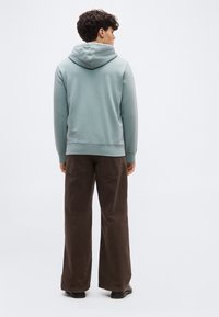 Νεαρός άνδρας με γυρισμένη πλάτη που φοράει γαλάζιο φούτερ με κουκούλα, φαρδιά καφέ παντελόνια και μαύρα παπούτσια μπροστά σε λευκό, απλό φόντο.