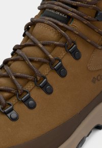 Gros plan d'une botte de randonnée en daim marron avec des lacets marron foncé passés à travers des œillets métalliques noirs.