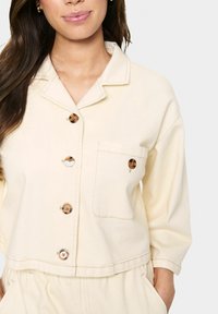 Giacca button-up corta color crema, con colletto, due tasche a toppa e bottoni marroni; realizzata in un tessuto morbido e texturizzato.