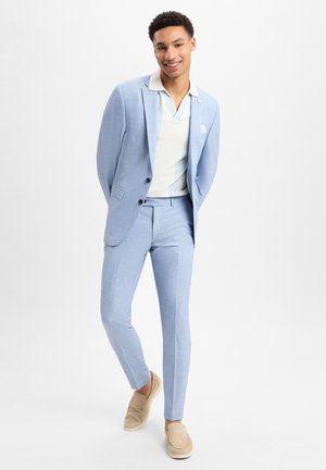 Hellblauer Anzug aus strukturiertem Stoff, kombiniert mit einem cremefarbenen Poloshirt mit blauen Streifen. Beigefarbene Slipper runden das Outfit ab.