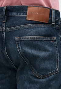 Tumesinised teksapüksid, millel on pruun nahast plaat "TOMMY HILFIGER" sildiga, nähtav õmblus ja minimalistliku disainiga tagataskud.