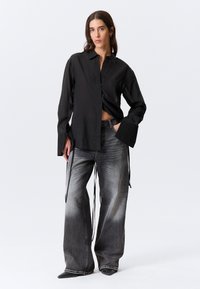 Chemise noire à boutons avec larges poignets, associée à un jean gris délavé ample avec cordons de serrage. Le mannequin est placé devant un fond uni.