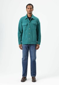 Turfkleurig, zwaar button-up jasje met twee borstzakken, gedragen over een zwart shirt, gecombineerd met blauwe spijkerbroek en bruine schoenen.