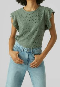T-shirt à manches courtes vert olive avec motif en œillets et manches volantes, associé à un jean taille haute bleu clair.