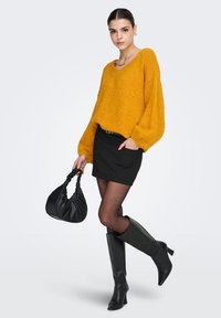 Sennepsgul strikkegenser med vide ermer, svart miniskjørt, gjennomsiktige svarte strømpebukser, svarte knelang støvler og en svart veske.