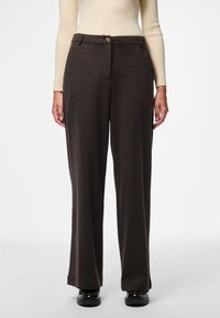 Kobieta nosząca ciemnobrązowe szerokie spodnie, beżowy prążkowany top z długim rękawem i czarne błyszczące buty na białym tle.