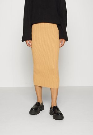 Person iført en sort sweater, ribbet kamelfarvet midi nederdel og sorte chunky skinnende loafers, stående mod en ensartet lys baggrund.