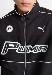 Svart glidelåsjakke med hvite detaljer, iøynefallende "PUMA"-logo og en sirkulær "P"-logo. Glatt stoff med høy krage og glidelås.