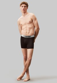 Boxers en coton noir avec une ceinture élastique grise affichant "Calvin Klein." Le modèle se tient pieds nus, mettant en valeur un design ajusté.