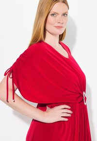 Punainen mekko, jossa on rento, valuva muotoilu, varustettuna kerrostetuilla hihoilla ja koristeellisella vyökoristeella vyötäröllä. Silkkinen, kiiltävä kankaan rakenne.