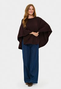 Temno rjava majica širokih rokavov z ohlapnim krojem, v kombinaciji z oversized modrimi jeans hlačami. Tkanina se zdi mehka in teksturirana z sproščenim dizajnom.