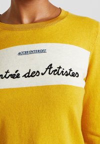 Żółty sweter z dzianiny z białym poziomym pasem, na którym wyszyty tekst: "ACCES INTERDIT" powyżej i "Entrée des Artistes" poniżej.