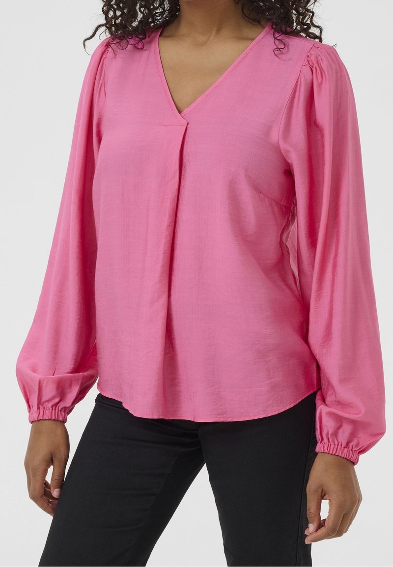 Blouse rose à manches longues avec un col en V, dotée d'épaules froncées et de poignets élastiques. Confectionnée dans un tissu léger et texturé.