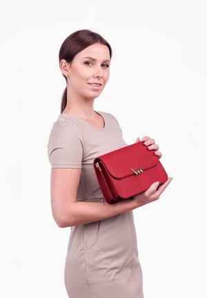 Červená kožená kabelka se zlatým akcentem ve tvaru písmene 'W'. Obsahuje klopu a má obdélníkový design, držená modelem.