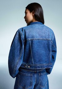 Traper jakna s teksturiranim rukavima, tamnoplave boje, skraćenog kroja i srebrnim detaljima. Sadrži karakteristične točkaste uzorke na rukavima.