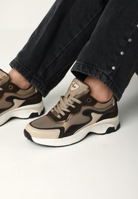 Zapatillas beige y marrón con paneles de malla y cuero, cordones con un acabado texturizado, acentos negros y diseño de suela blanca.