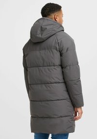 Grå oppustelig frakke med vandret kviltet design, hætte og lange ærmer. Materialet ser glat og let skinnende ud og strækker sig til midten af låret.