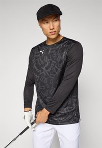 Svart langermet golf skjorte med et teksturert mønster, med en hvit logo på brystet. Kombinert med hvite bukser og en golfhanske.