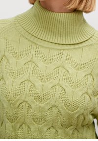 Helles grünes Kabelstrickpullover mit hohem geripptem Kragen. Auffällige Textur und Muster aus ineinander verwobenen Schlaufen. Weicher Stoff.