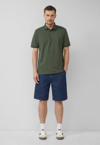 Groene polo met een marineblauwe kraag, gecombineerd met blauwe denim shorts. Het model draagt witte sokken en sneakers, staand tegen een neutrale achtergrond.