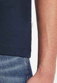 Donkerblauwe katoenen t-shirt met korte mouwen, voorzien van een wit logo etiket aan de zoom. Gecombineerd met vervaagde blauwe denim jeans.