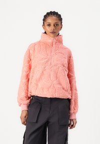 Korallifleece-pullover, jossa on korkeakaulus, vetoketjukiinnitys ja teksturoitu kuvio. Pitkät hihat, joissa on resorit, yhdistettynä tummiin cargo-housuihin.