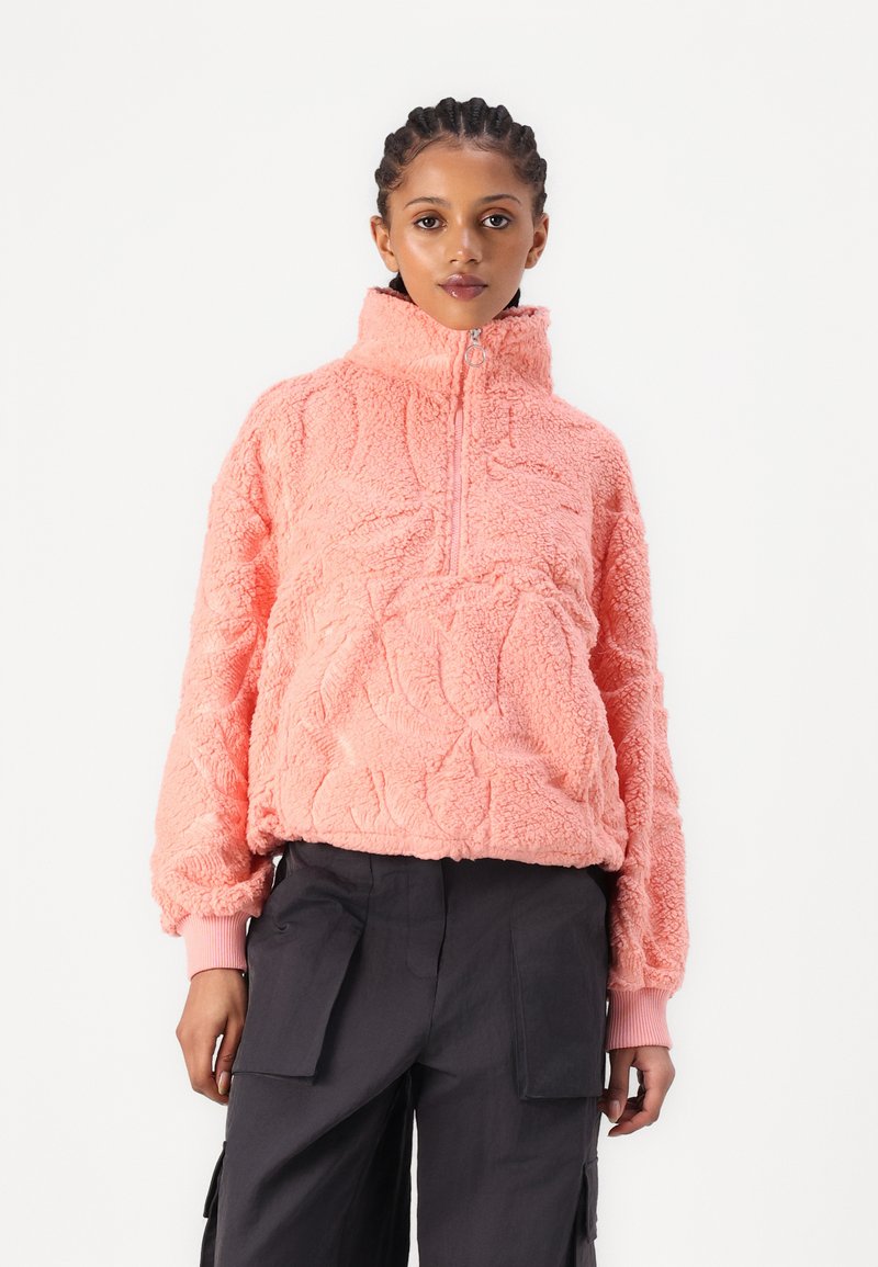 Korallifleece-pullover, jossa on korkeakaulus, vetoketjukiinnitys ja teksturoitu kuvio. Pitkät hihat, joissa on resorit, yhdistettynä tummiin cargo-housuihin.