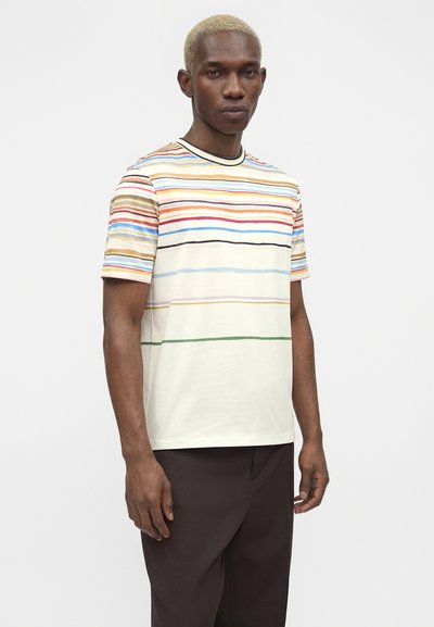 Άνδρας με κοντά ξανθά μαλλιά που φοράει λευκό μπλουζάκι με πολύχρωμες οριζόντιες ρίγες και σκούρο καφέ παντελόνι, στέκεται μπροστά σε απλό φόντο.