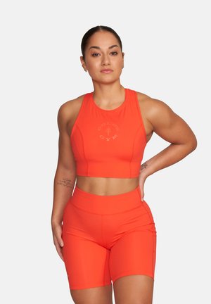Fit woman with hair in a bun wearing bright orange Gold's Gym crop top and matching high-waisted biker shorts, posing with hand on hip.