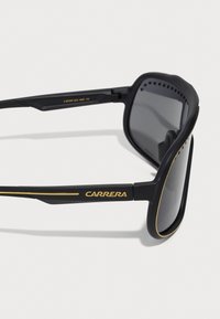 Lunettes de sport noires avec une finition mate, accents dorés sur les branches et verres teintés foncés. Présente des trous de ventilation le long de la monture.