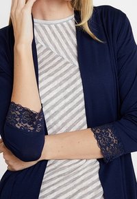 Mujer vestida con un cárdigan azul marino con puños de encaje sobre una camiseta de rayas grises y blancas, mostrada desde los hombros hasta la cintura.