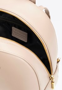 Zaino in pelle beige con forma rotonda, zip dorata e interno nero con etichetta del logo. Include una piccola tasca con zip all'interno.