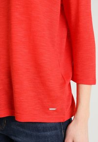 Pull texturé rouge à manches trois-quarts, doté d'une petite étiquette logo en métal en bas à gauche et d'une coupe décontractée.