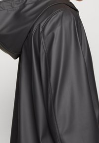 Veste imperméable noire avec capuche, pr ésentant une texture lisse et brillante ainsi que des coutures contrastantes. Le design comprend une coupe décontractée et des épaules tombantes.