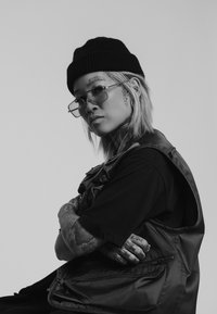 Person som bär en svart mössa och solglasögon, med synliga tatueringar, lager-på-lager med en mörk väst över en svart skjorta. Monokrom bakgrund.