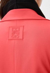 Różowy koralowy płaszcz z teksturowanej tkaniny, z czarnym kołnierzem i wypukłym, gumowym logo w górnej części pleców.