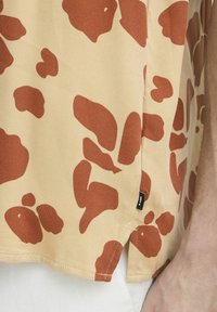 Camisa de manga corta en un tejido marrón claro que presenta un gran patrón abstracto en marrón rojizo, con un detalle de etiqueta en la costura lateral. El dobladillo es ligeramente curvado.