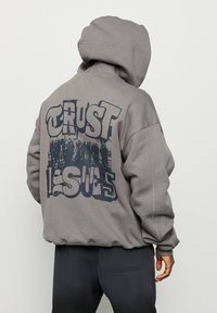 Sweat à capuche gris avec cordon de serrage, présentant une grande impression graphique au dos avec l'inscription "TRUST ISSUES". Texture douce et coupe décontractée.