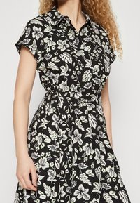 Blommig skjortklänning i svart tyg med vita och ljusgröna blad- och blommönster, med krage och bälte i midjan.