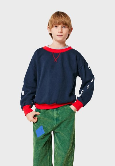 Felpa navy con accenti rossi, polsini a coste e grandi lettere bianche sulle maniche, abbinata a pantaloni in velluto a coste verdi con una toppa blu.