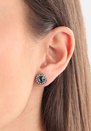 Pendientes de botón de gema negra rodeados de cristales transparentes, engastados en un tono plateado circular. Primer plano junto a una oreja con cabello lacio.