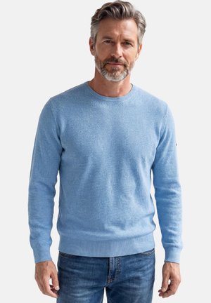 Mand i midten af 50'erne med gråt hår og skæg iført en lyseblå tekstureret sweater og blå jeans, stående foran en ensfarvet baggrund.