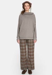 Jasnoszara oversizeowa bluza z kapturem typu komin i kieszenią z przodu, zestawiona z szerokimi brązowymi spodniami w kratę. Dopełnieniem stroju są brązowe buty.