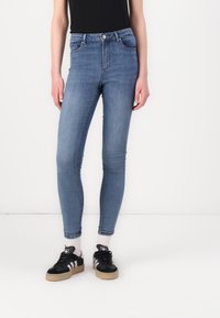 Jeanși skinny cu talie înaltă, de culoare albastră, realizați din denim, cu cinci buzunare, ușor decolorati și o siluetă ajustată, asortați cu adidași negri.