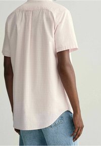 Camisa de manga corta en tejido de color rosa claro con un patrón de cuadros, con un dobladillo recto y detalle en el yugo trasero. Combinada con jeans de color azul claro.