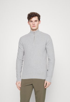 Un pull zippé gris clair en maille texturée avec un col haut et une fermeture éclair argentée, associé à un pantalon vert olive.