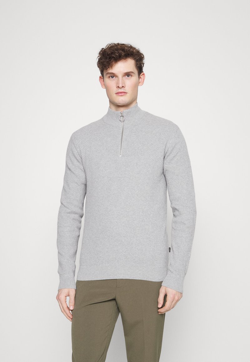 Un pull zippé gris clair en maille texturée avec un col haut et une fermeture éclair argentée, associé à un pantalon vert olive.