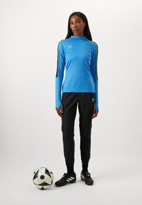 Sporta apģērbs, kurā ir zils garām piedurknēm tops ar melniem un dzelteniem akcentiem, saskaņots ar melnām biksēm un melniem futbola apaviem. Blakus ir futbola bumba.