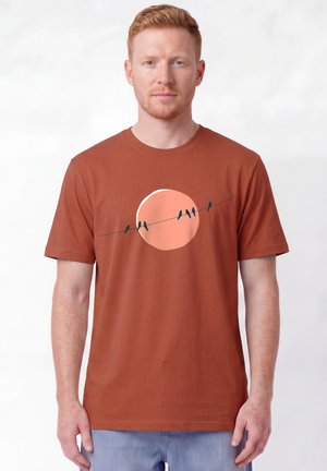 Homme aux cheveux roux portant un t-shirt couleur rouille avec un grand cercle rose et cinq petits oiseaux assis sur un fil.