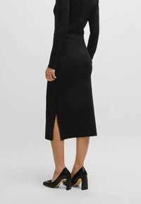 Jupe midi tricotée noire avec une fente latérale, texture lisse, design ajusté et taille haute, associée à des chaussures à talons hauts noires.